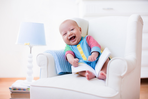Cute funny baby boy reading a book sitting in a white chair at home. Children read books in a library seat. Nursery and playroom interior for kids. Early development and learning for young kid.